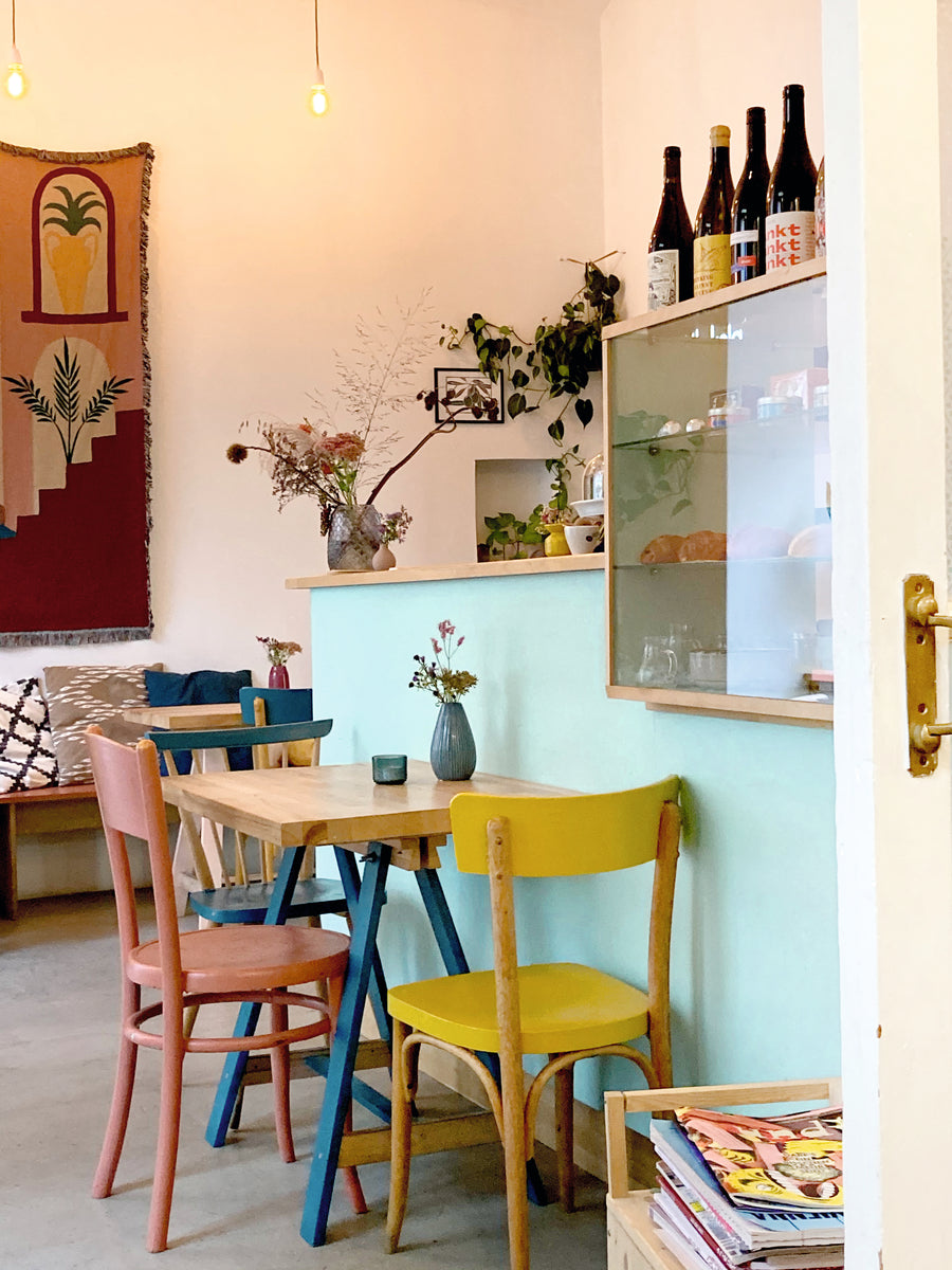 sous-bois Café-Bar mit pastelfarbenen Sesseln, gemütlichen Kissen und begrünten Wänden. Getränke und Gebäck sind in der Auslage.